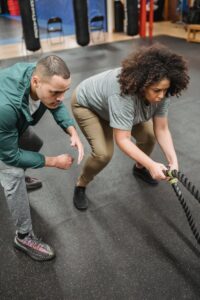 From above of concentrated young male trainer instructing determined obese African American woman doing exercise on battle ropes during functional workout in gym with