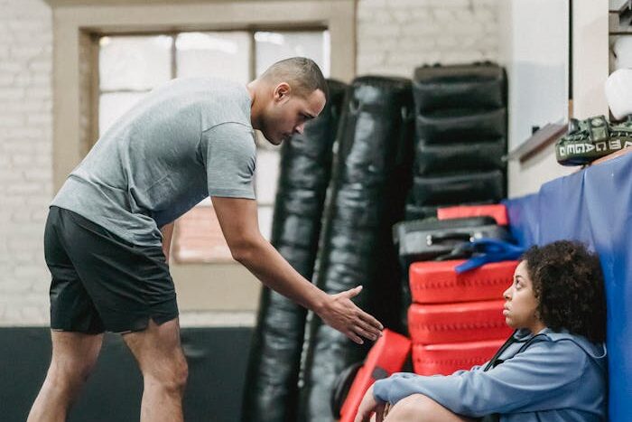 Full body side view of muscular black personal coach reaching out hand to African American female while training in gym